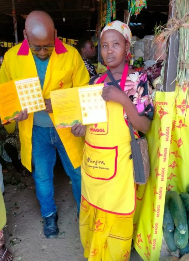 A vendor in a MAGGI apron stands beside her branded table top, holding a MAGGI programme card